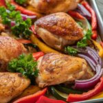 Sheet pan Greek chicken roasted with bell peppers, red onion, and zucchini, garnished with fresh parsley and lemon